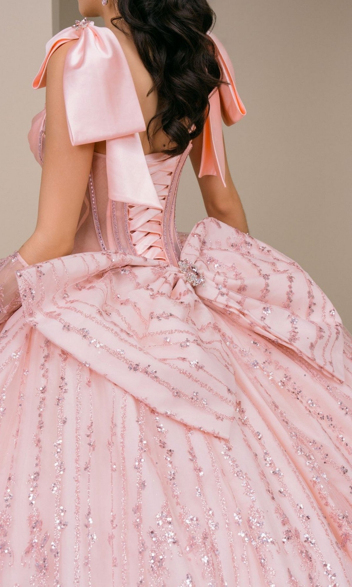 A woman is shown from behind wearing the Quinceanera Dress 8530JQ by Cinderella Couture, a pastel pink ball gown adorned with sequin details and a large bow at the back, featuring a stunning beaded corset bodice.